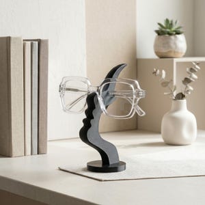 May include: A decorative eyeglasses holder in the shape of a stylized face, in black and gray. Clear-framed eyeglasses rest on the holder. The holder is on a white surface, with books and a small plant in the background.