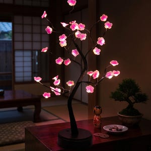May include: A decorative cherry blossom tree lamp with pink illuminated flowers and a dark brown trunk. The lamp is on a wooden table, with a bonsai tree and Japanese doll in the background. The lamp is a unique home decor item.