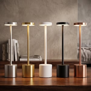 May include: Five contemporary table lamps in silver, gold, white, black, and copper. Each lamp has a cylindrical base, a slender stem, and a round light. The lamps are displayed on a dark wooden surface.
