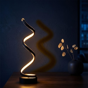 May include: A contemporary black and gold spiral LED table lamp. The lamp features a curved, ribbon-like design with a warm white light. The lamp sits on a round black base. A vase with gold-coloured leaves is in the background.
