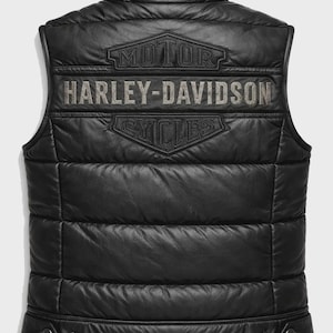 Könnte beinhalten: Eine schwarze, gesteppte Lederweste mit Stehkragen und horizontalen Nähten. Auf der Rückseite befindet sich ein geprägtes Harley-Davidson Motor Cycles Logo mit dem Schriftzug Harley-Davidson in hellgrauer Farbe im oberen Bereich.