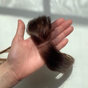 Puede incluir: Un mechón de extensiones de cabello castaño oscuro, sostenido en una mano. El cabello presenta una mezcla de tonos marrones claros y oscuros, fijado a una base fina y enrejada. Las extensiones parecen nuevas y sin usar.