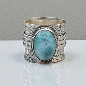May include: A silver ring with an oval, light blue gemstone. The ring has a wide band with floral patterns and three horizontal bands. The gemstone is set in a silver bezel.
