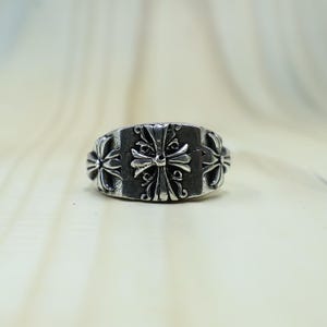Könnte beinhalten: Ein silberfarbener Metallring mit einem breiten Band und einer dunklen, oxidierten Oberfläche. Das Design zeigt drei geprägte Kreuzmotive, wobei ein größeres zentrales Kreuz von zwei kleineren, identischen Kreuzen flankiert wird.
