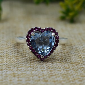 May include: A heart-shaped ring featuring a light blue gemstone center, surrounded by smaller, deep red gemstones. The ring has a silver band and is set against a neutral background.