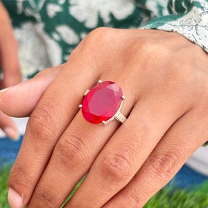 Puede incluir: Un anillo de plata con una gran piedra preciosa roja rubí de talla ovalada. El anillo tiene una banda sencilla y está engastado con seis garras. La piedra preciosa está facetada, captando la luz y mostrando un color rojo vibrante. El anillo es una pieza llamativa.