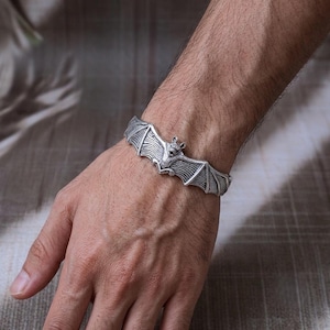 May include: A silver-toned metal cuff bracelet featuring a detailed bat design with outstretched wings. The bat is positioned in the center, with its wings extending along the band, showcasing textured lines and defined features.
