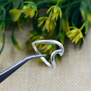 May include: A silver ring with a distinctive wave design. The ring is held by tweezers against a backdrop of green and yellow foliage. The ring's design features a curved, open shape.