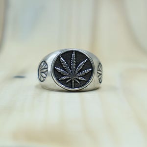 May include: A silver-toned metal ring featuring a circular centerpiece with a raised, detailed cannabis leaf design set against a dark, recessed background. The sides of the ring band are decorated with smaller, engraved sunburst or leaf-like motifs.