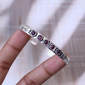 May include: A silver-toned bracelet with a series of round, deep red gemstones. The bracelet has a geometric design with triangles and curved shapes. The gemstones are evenly spaced along the bracelet's surface, creating a symmetrical pattern.