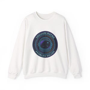 May include: White crewneck sweatshirt with a circular graphic featuring an astronaut helmet, stars, and the text "VACUUM-RATED PROBLEM SOLVER" in a blue and teal design. The sweatshirt is made of a soft material.