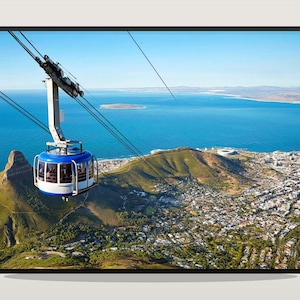 May include: A scenic aerial view of a city and coastline, with a blue and white cable car ascending a cable. The cable car is suspended above a green mountain and the city below. The sky is clear blue.