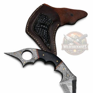 May include: A Damascus steel knife with a curved blade and a black handle with gold accents. The knife is paired with a brown leather sheath. The blade has a unique pattern, and the handle has finger grooves for a secure grip. The knife is a hunting or utility tool.