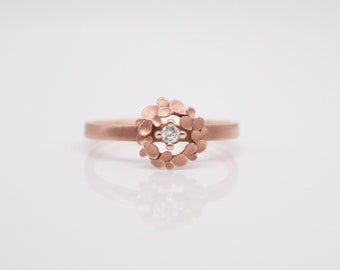 May include: A rose gold ring with a delicate diamond set in a center surrounded by a ring of small, textured, rose gold leaves.