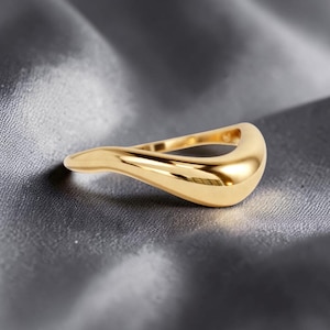 May include: A gold-colored ring with a smooth, flowing design. The ring has a curved shape with a rounded top and a slender band. The polished surface reflects light, creating a bright, shiny appearance. The ring is likely made of gold or a gold-colored metal.