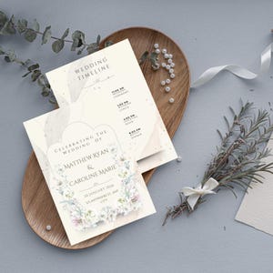 May include: Wedding invitation and timeline cards with floral accents. The invitation reads "Celebrating the Wedding of Matthew Ryan & Caroline Marie" with the date and location. The timeline card lists the ceremony, lunch, cocktail hour, and dinner times.
