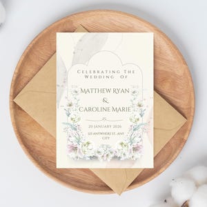 Peut inclure: Une invitation de mariage avec des motifs floraux, avec les noms Matthew Ryan et Caroline Marie. La carte est sur une assiette en bois avec une enveloppe marron. Le texte dit "Célébrant le mariage de" et la date est le 20 janvier 2026.