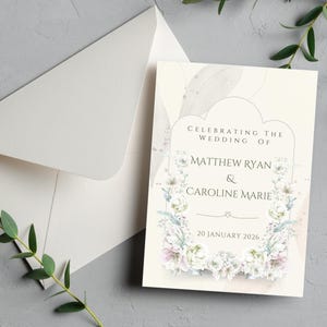 Puede incluir: Una tarjeta de invitación de boda crema con ilustraciones florales y el texto "Celebrating the Wedding of Matthew Ryan & Caroline Marie" y la fecha "20 January 2026". También hay un sobre blanco.