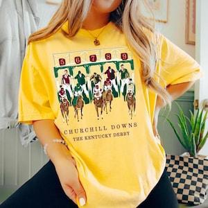 May include: Yellow t-shirt featuring a graphic of a horse race at Churchill Downs, with the numbers 5 through 9 above the track. The text "CHURCHILL DOWNS THE KENTUCKY DERBY" is printed below the image.