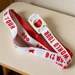 May include: A red and white lanyard with the text "WORLD TOUR" in red capital letters. The lanyard has a white plastic clasp and features additional red text and graphics. The lanyard is laying on a light-colored surface.