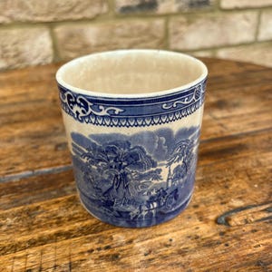 May include: A ceramic cup with a blue and white landscape design. The cup features a detailed illustration of trees, buildings, and figures. The rim has a decorative blue border. The cup is on a wooden surface.