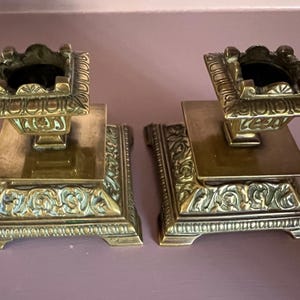 May include: A pair of matching square brass candle holders featuring ornate relief detailing on the base and a decorative rim around the top opening. The metal has a polished finish with darkened recessed areas highlighting the intricate patterns.