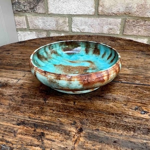 May include: A handmade ceramic bowl with a turquoise and brown glazed interior. The exterior has a band of brown and turquoise, creating a rustic, artistic aesthetic. The bowl is displayed on a wooden surface.