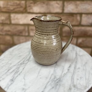 May include: A ceramic jug with a speckled cream-coloured glaze and horizontal ribbed texture. The vessel features a wide, rounded body, a flared rim with a pouring spout, and a single curved handle with a dark brown rim accent.