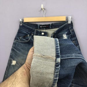 May include: Blue denim jeans with distressed details, hanging on a wooden hanger. The jeans have a classic five-pocket design and a faded wash. The cuff of the jeans is turned up, revealing the lighter-colored interior fabric.