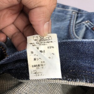 May include: Close-up of a blue denim garment with a white tag. The tag displays washing instructions, including a 40-degree wash symbol, and Japanese text indicating fabric composition and care details. The fabric is 93% cotton.
