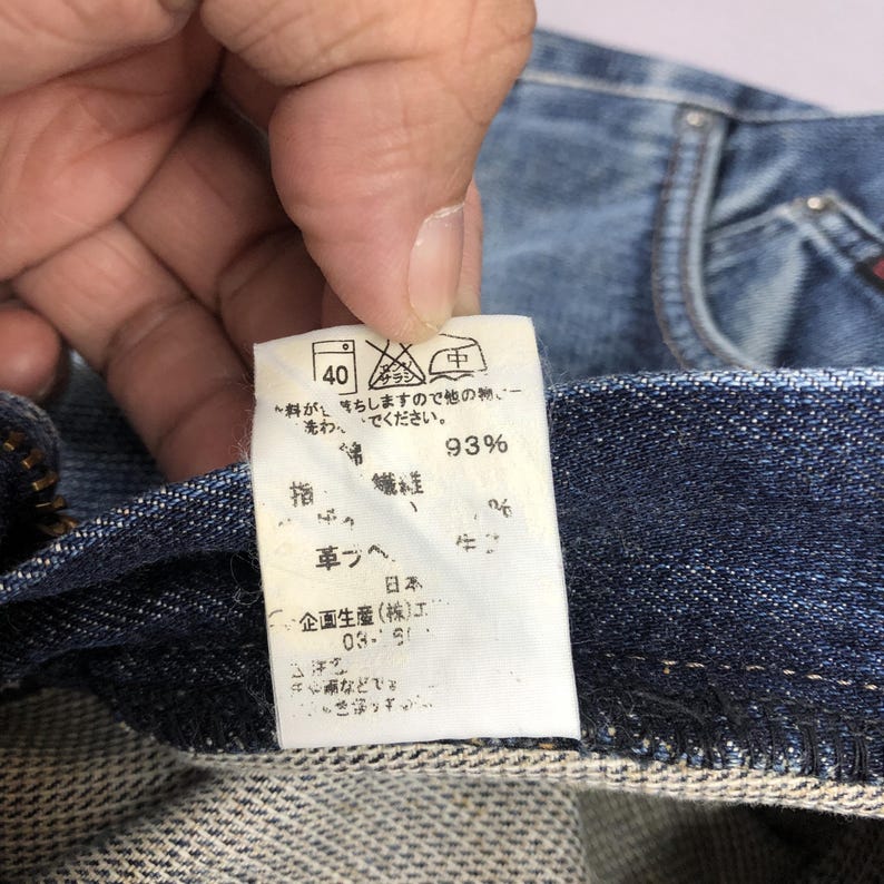 May include: Close-up of a blue denim garment with a white tag. The tag displays washing instructions, including a 40-degree wash symbol, and Japanese text indicating fabric composition and care details. The fabric is 93% cotton.
