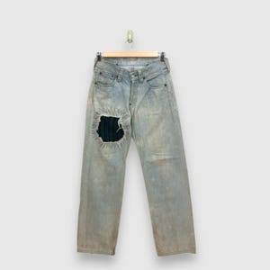 May include: Light-wash blue denim jeans featuring a distressed finish and a prominent rectangular patch on the left thigh. The patch is made of a dark green, blue, and black striped fabric, secured with visible, decorative white stitching.