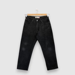 Puede incluir: Unos vaqueros de mezclilla negra con corte relajado, bragueta de botones, cinco bolsillos y una etiqueta interior visible con la inscripción RELAXED FIT. Los vaqueros están colgados en una percha de madera y presentan un lavado desgastado.