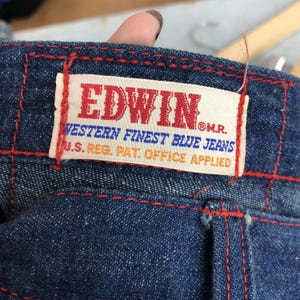 Puede incluir: Primer plano de unos vaqueros azules con costuras rojas. Una etiqueta rectangular con la marca "EDWIN" en rojo est&aacute; cosida a los vaqueros. La etiqueta tambi&eacute;n incluye el texto "WESTERN FINEST BLUE JEANS" y "U.S. REG. PAT. OFFICE APPLIED".
