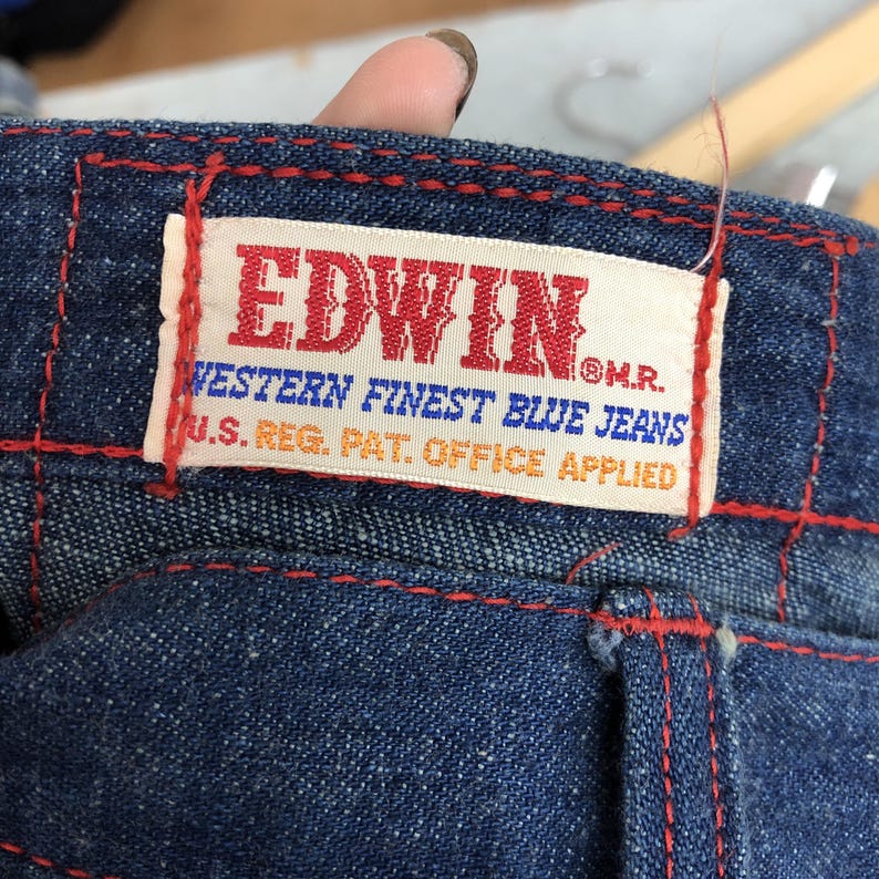 Puede incluir: Primer plano de unos vaqueros azules con costuras rojas. Una etiqueta rectangular con la marca "EDWIN" en rojo est&aacute; cosida a los vaqueros. La etiqueta tambi&eacute;n incluye el texto "WESTERN FINEST BLUE JEANS" y "U.S. REG. PAT. OFFICE APPLIED".