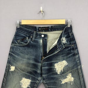 May include: Distressed blue denim jeans with multiple ripped sections. The jeans are hanging on a wooden hanger against a gray background. The zipper is partially open, and the jeans have a faded appearance.