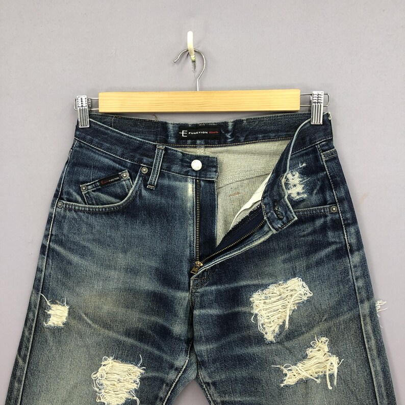 May include: Distressed blue denim jeans with multiple ripped sections. The jeans are hanging on a wooden hanger against a gray background. The zipper is partially open, and the jeans have a faded appearance.