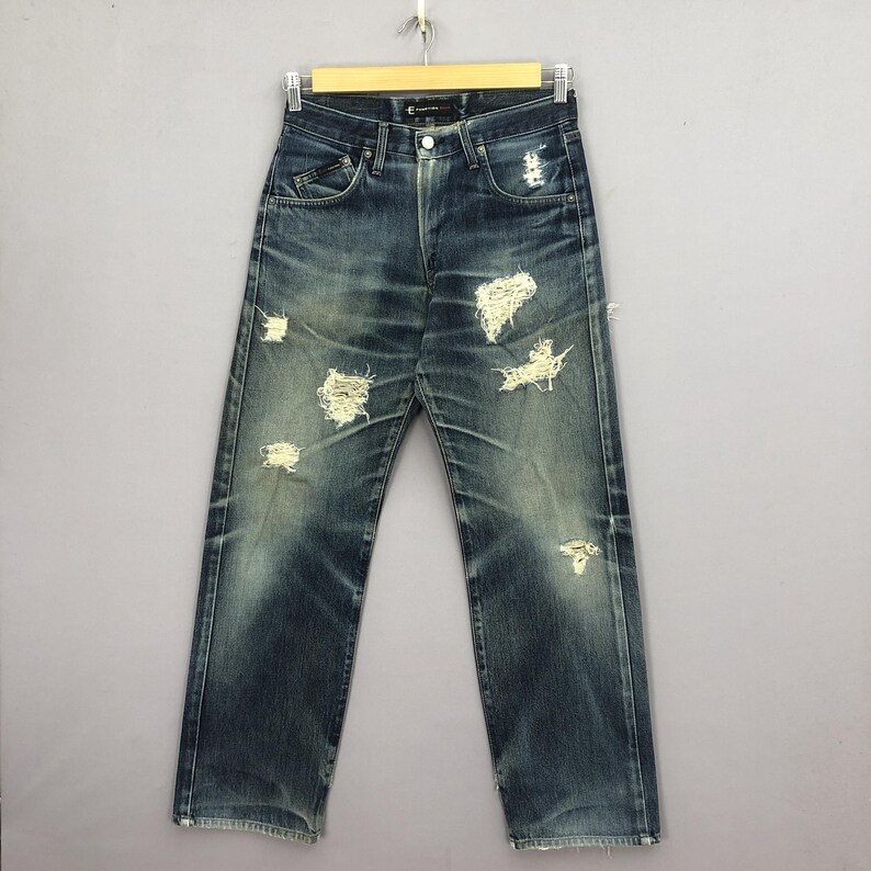 May include: Blue denim jeans with distressed details, including multiple rips and faded areas. The jeans are displayed on a hanger against a neutral background, showcasing a classic five-pocket design.