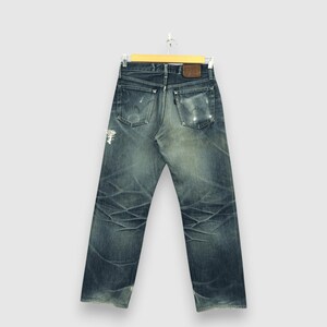 May include: Vintage blue denim jeans with distressed details, including faded areas and rips. The jeans are hung on a wooden hanger, showcasing the back pockets and a leather patch. The denim has a worn, textured appearance.