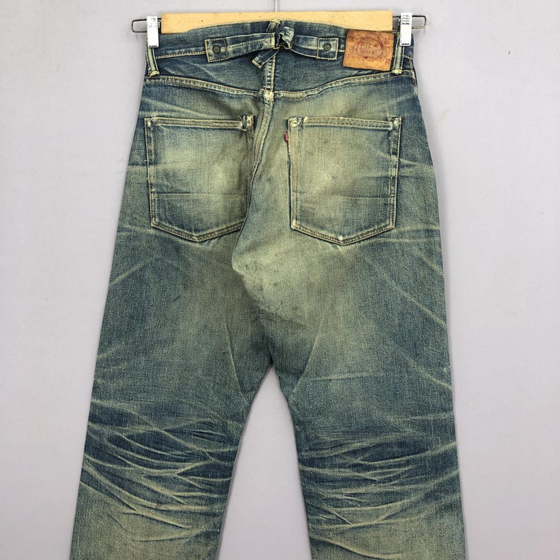 Puede incluir: Vaqueros vintage de mezclilla azul con un aspecto desgastado y envejecido. Los vaqueros tienen dos bolsillos traseros, un parche de cuero y cierre de botones. La mezclilla presenta zonas descoloridas y marcas de desgaste visibles.
