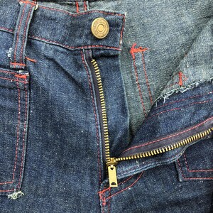 Puede incluir: Primer plano de unos vaqueros de mezclilla azul oscuro con una cremallera de lat&oacute;n parcialmente desabrochada. Las costuras rojas acent&uacute;an las costuras y los bolsillos. Se ve un bot&oacute;n de lat&oacute;n en la cintura. Los vaqueros parecen desgastados.