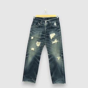 May include: Blue denim jeans with distressed details, including ripped sections. The jeans are hanging on a wooden hanger against a light background. The jeans have a classic five-pocket design.