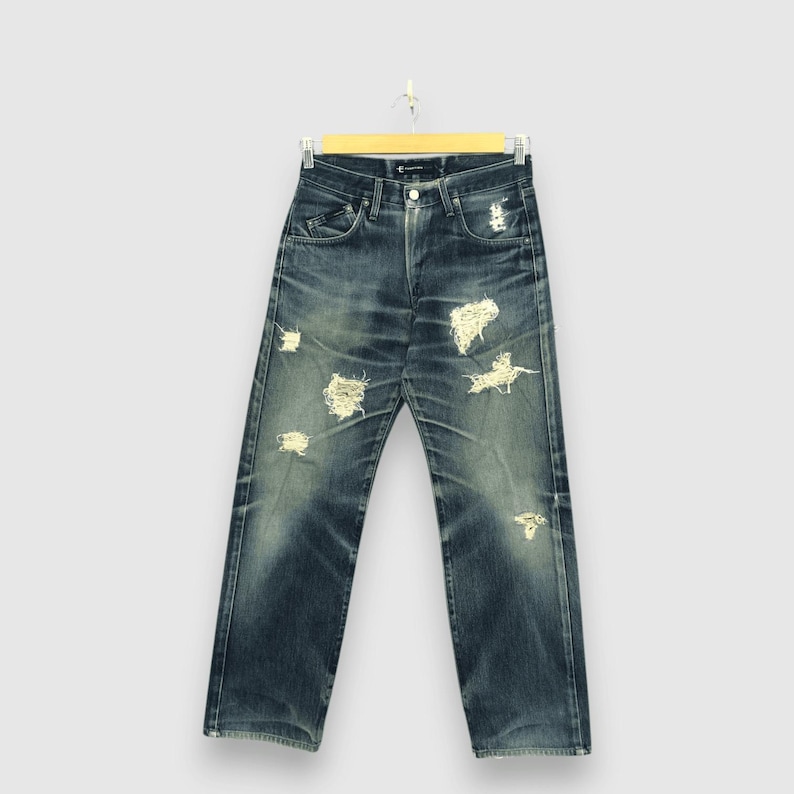 May include: Blue denim jeans with distressed details, including ripped sections. The jeans are hanging on a wooden hanger against a light background. The jeans have a classic five-pocket design.