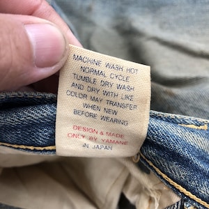 Puede incluir: Primer plano de una etiqueta de vaqueros de mezclilla con instrucciones de cuidado. La etiqueta es beige con texto negro: "MACHINE WASH HOT, TUMBLE DRY WASH, DESIGN & MADE ONLY BY YAMANE IN JAPAN."
