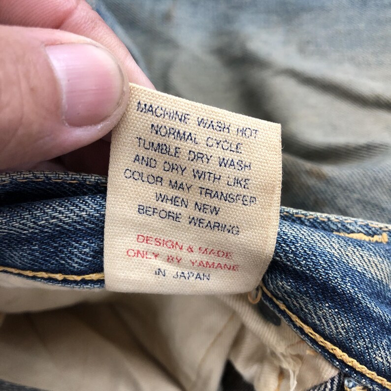 Puede incluir: Primer plano de una etiqueta de vaqueros de mezclilla con instrucciones de cuidado. La etiqueta es beige con texto negro: "MACHINE WASH HOT, TUMBLE DRY WASH, DESIGN & MADE ONLY BY YAMANE IN JAPAN."