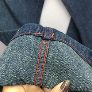 Puede incluir: Primer plano de unos vaqueros de mezclilla azules con costuras naranjas. Los vaqueros muestran los detalles del dobladillo y las costuras, destacando el contraste entre la mezclilla y las costuras brillantes. La tela tiene un aspecto texturizado.