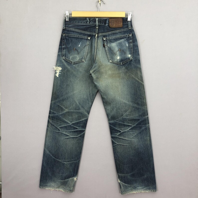 May include: Faded blue denim jeans with distressed details, including rips and wear marks. The jeans have a classic five-pocket design, a brown leather patch, and are hanging on a wooden hanger.