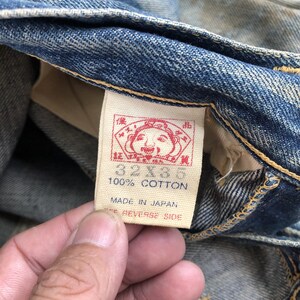 Puede incluir: Primer plano de una etiqueta de jeans de mezclilla. La etiqueta es rectangular con un gr&aacute;fico rojo y dorado y el texto "32x35" y "100% Algod&oacute;n". La etiqueta tambi&eacute;n indica "Made in Japan".