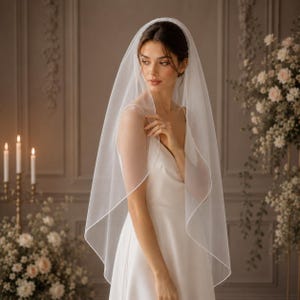May include: A bride wearing a white wedding veil with a delicate, thin trim. The veil is draped over the head and shoulders, complementing a white wedding dress. The background features floral arrangements and candles, creating a romantic atmosphere.