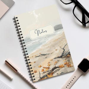 Könnte beinhalten: Ein spiralgebundenes Notizbuch mit einer gedruckten Illustration eines Sandstrandes mit verstreuten Herbstblättern und Treibholz. Der Einband zeigt ein cremefarbenes rechteckiges Etikett mit dem Wort Notes in dunkelblauer Schreibschrift.